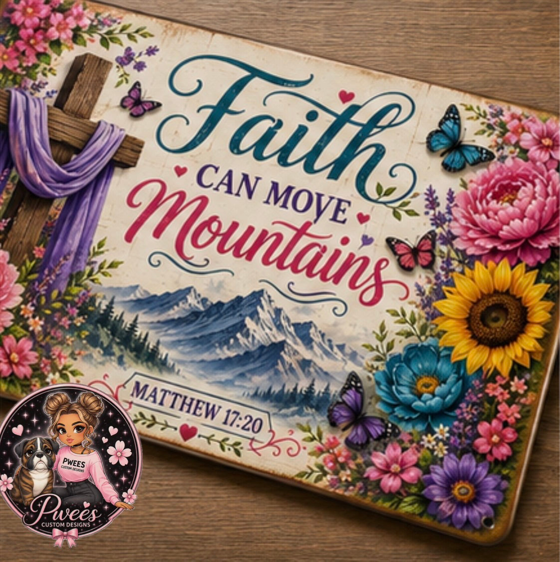 A decorative rectangular sign with the phrase “Faith can move mountains” and “Matthew 17:20,” featuring a mountain landscape illustration, colorful flowers, butterflies, and a wooden cross with a purple scarf on a wood surface. A circular watermark/logo appears in the lower left corner.