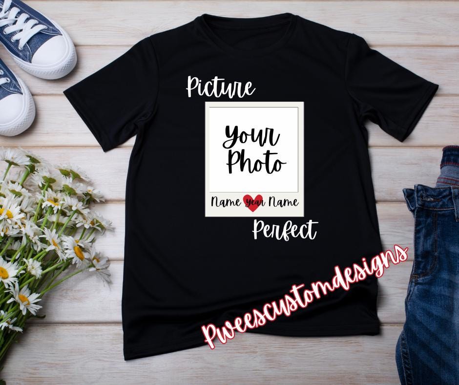 Black t-shirt with customizable text design on a wooden surface with shoes and flowers.