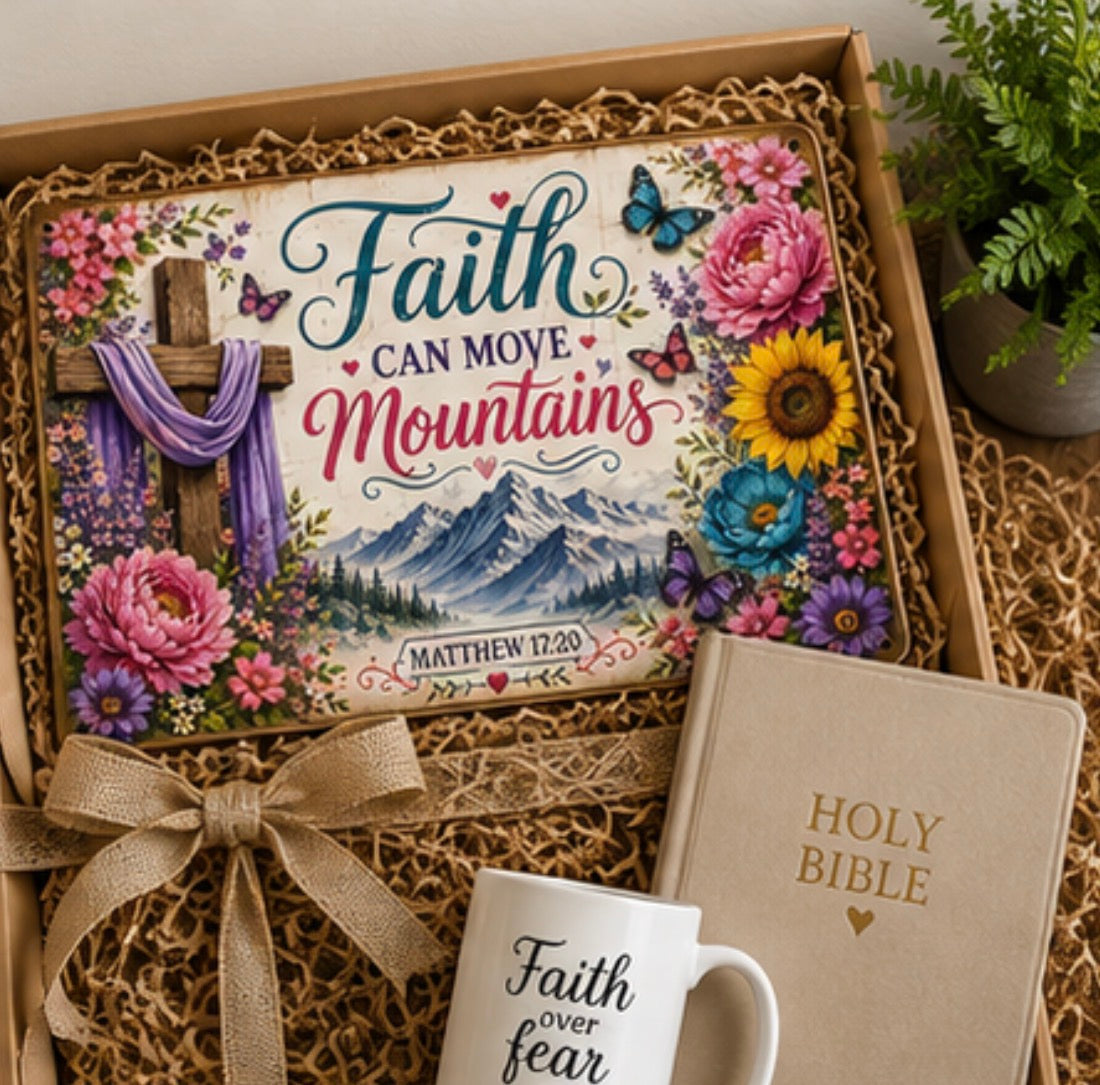 A gift box arrangement featuring a decorative print with the words 'Faith can move mountains,' a cross, flowers, butterflies, and a mountain scene, styled with a beige Bible, a white mug reading 'Faith over fear,' and a ribbon bow on a neutral surface.