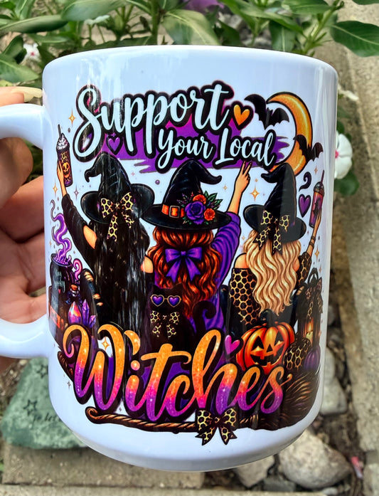A white ceramic mug held by a hand, featuring a colorful Halloween graphic with three witches, pumpkins, bats, and the text 'Support your local Witches'.