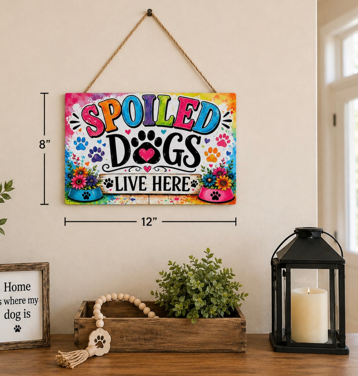 A colorful hanging wall sign that reads “SPOILED DOGS LIVE HERE” with paw prints and flowers, shown mounted on a beige wall above a wooden surface with decor items. Black dimension lines indicate 8 inches tall by 12 inches wide.