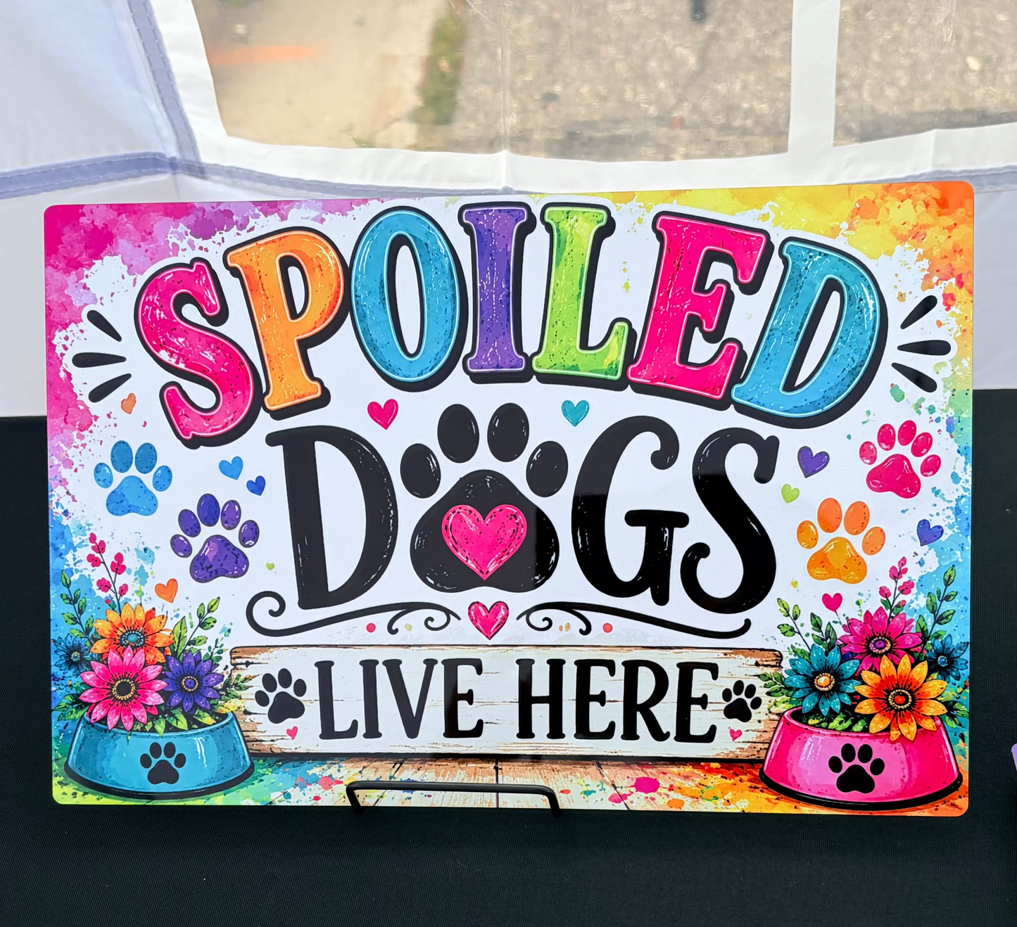 A colorful decorative sign that reads 'SPOILED DOGS LIVE HERE' with paw prints, flowers, and pet food bowls, displayed on a black surface.